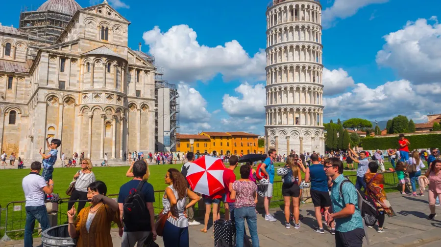 Tourists crowd Pisa’s Piazza dei Miracoli, posing playfully with Leaning Tower beside ornate Romanesque cathedral.