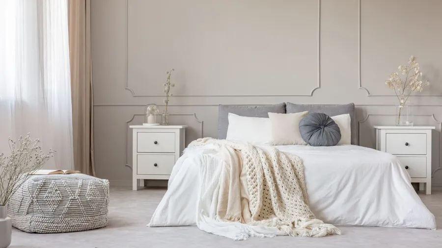A cozy, minimalist bedroom with a neatly made bed, white bedding, and decorative pillows, flanked by two white nightstands with vases and lamps, and a woven pouf on the left side.