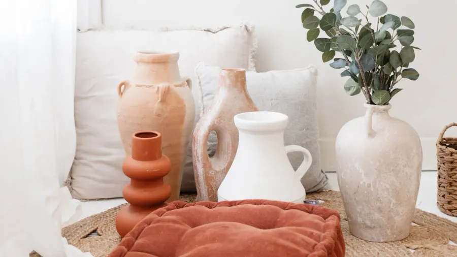 A collection of decorative vases and pottery in earthy tones, including terracotta and cream, arranged on a woven mat with soft cushions in the background.