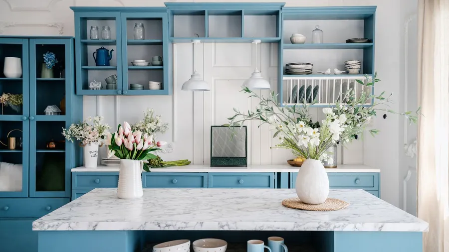 A stylish blue-and-white kitchen with open shelving and a marble island adorned with faux ceramic vases filled with white flowers and pink tulips.