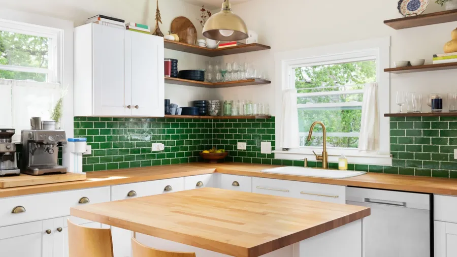 A kitchen interior with butcher block wooden countertops, white cabinets, open wooden shelves with dinnerware and jars, a gold fixture hanging over the island, and green subway tile backsplash.