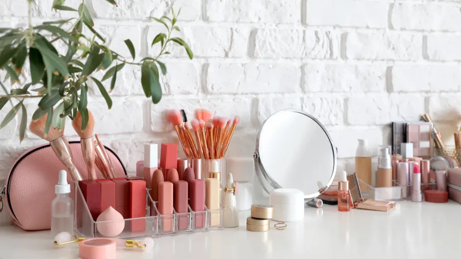 Neatly organized makeup in clear storage container and a small round mirror on a white dresser.