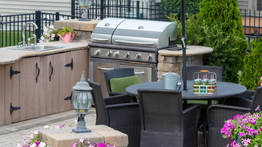 An outdoor kitchen on a patio with a built in grill, sink, wooden cabinets, and a sitting area with wicker table and chairs.