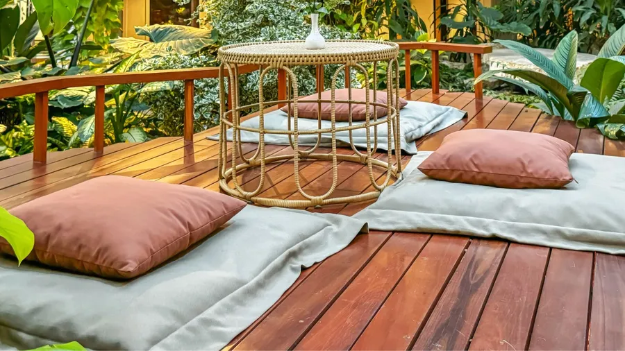 A wooden deck with cushions and a wicker table, surrounded by lush greenery.
