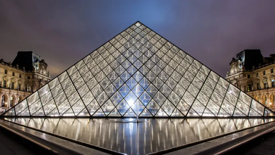 Night view of the illuminated glass pyramid entrance of the Louvre Museum in Paris, reflecting in the surrounding water with historic buildings in the background.
