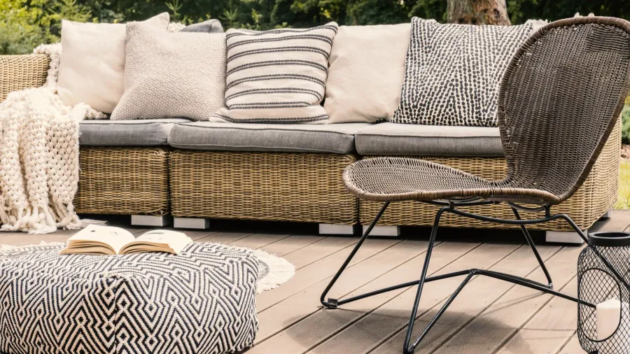 A stylish patio with rattan sofa with throw blanket and pillows, metal chair, and pouf with a book on a wooden floor.