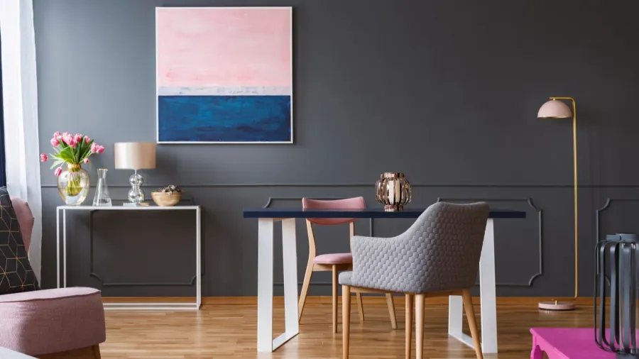 A modern dining area featuring a dark gray wall, a blue tabletop, pink chairs, and a colorful abstract painting.