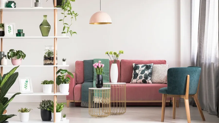 A living room interior with pink sofa with cushions and throw blankets, green armchair, shelf with decor elements, and coffee tables with vases.