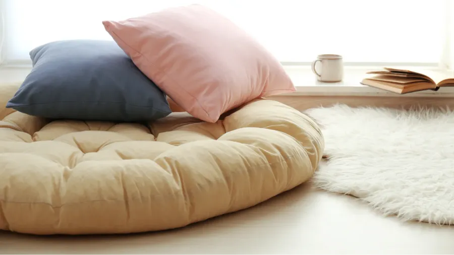 A floor cushion, pillows, and a faux fur rug near a window.