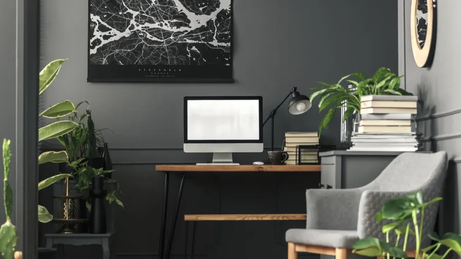 A home office interior with a gray armchair, wooden desk with books, computer, and lamp, and abstract artwork on the wall.