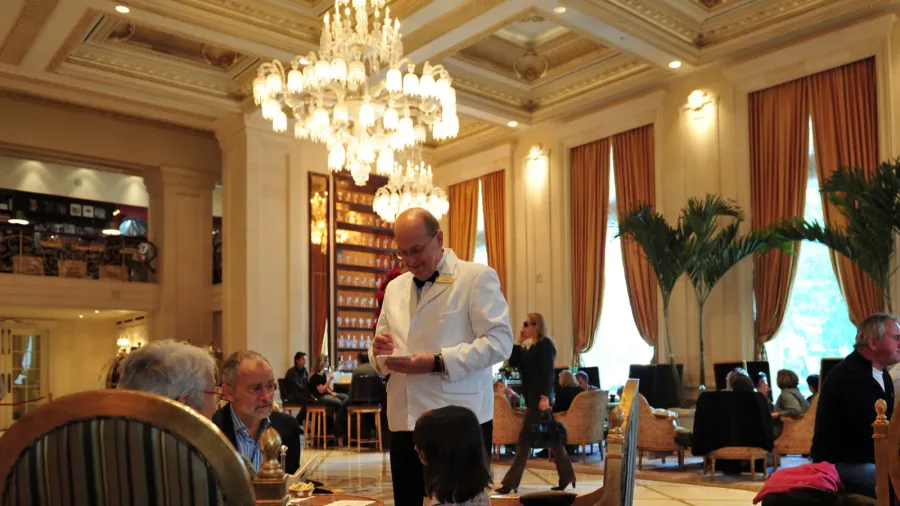 Restaurant in the Plaza Hotel featuring crystal chandelier, wall sconces, floor to ceiling windows with curtains, filled with people enjoying their meals and servers.