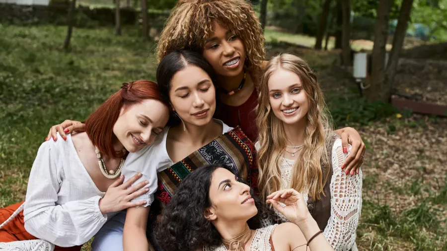 positive multiethnic women in boho outfits hugging while sitting on