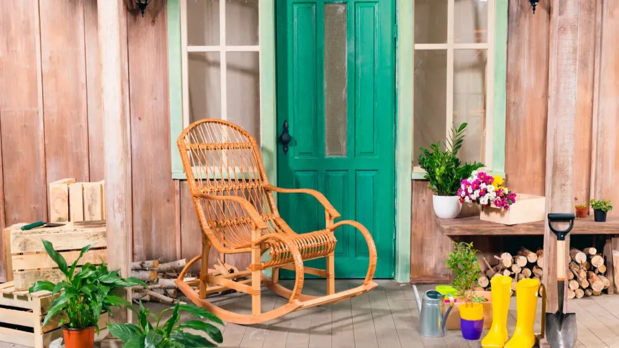 A wicker rocking chair in front of a bright green door with potted plants.