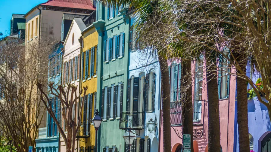 A row of historic, colorful houses known as Rainbow Row in Charleston, South Carolina, with palm trees and bare trees in the foreground.