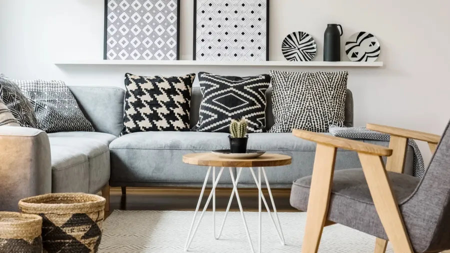 A living room interior featuring a gray sofa with black and white textured throw pillows, a gray armchair, white rug with coffee table on it, and black and white patterned art in frames and decor accents.