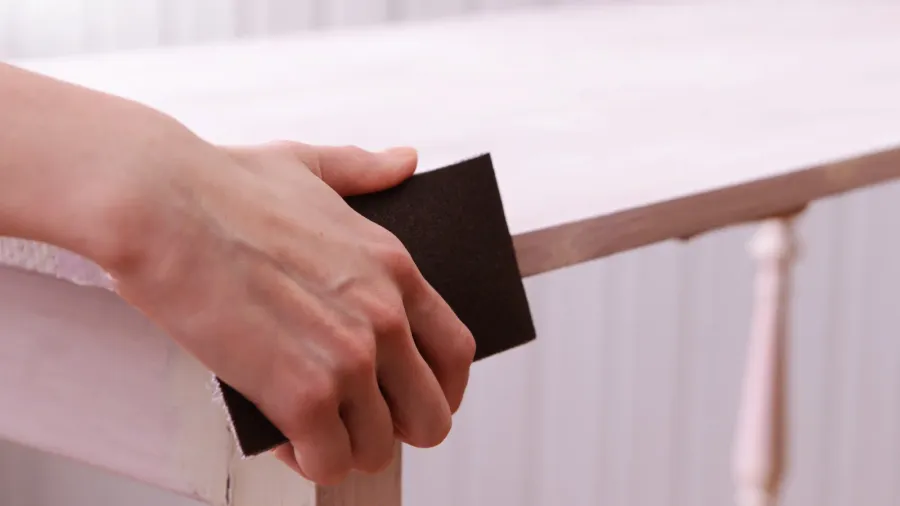 Closeup of a man's hand holding sand paper and sanding the edges of a wooden table.
