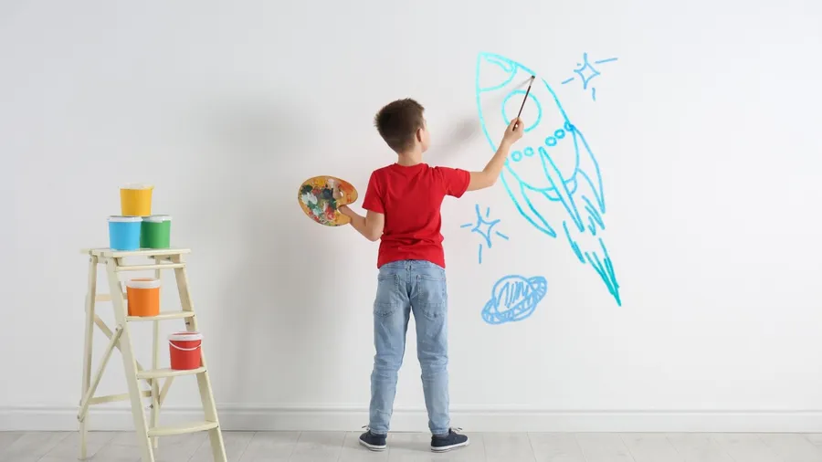 A child drawing a rocket with paints on a white wall.