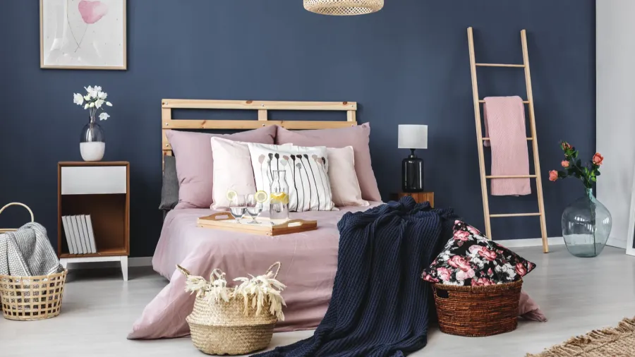 A cozy bedroom decorated with pastel bedding, storage baskets, and flowers, featuring a tray with drinks on the bed.
