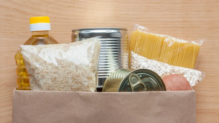 Pasta, rice, oatmeal, oil, potatoes and canned food in paper bag on wooden background top view.