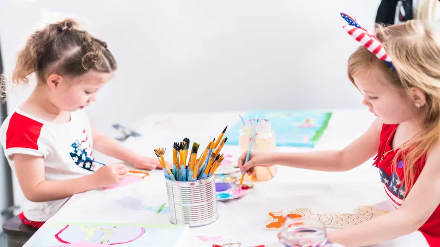 A crafts station with girls painting at a table with various brushes and supplies.