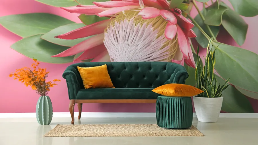 A living room interior with a velvet green sofa, flower vase, pillows, a plant pot, and floral wallpaper on the wall.