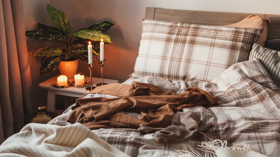 A cozy bedroom with flannel bedding, comforter, throw blanket, and burning candles and potted plant on the bedside table.