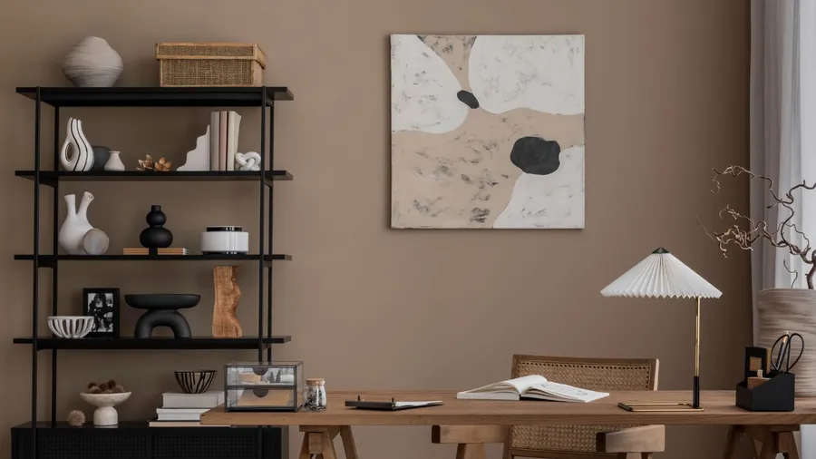 Aesthetic composition of living room interior with mock up poster frame, wooden desk, rattan chair, black rack, vase with branch, bookshelf with sculptural pieces and books, brown wall and office accessories. Home decor.