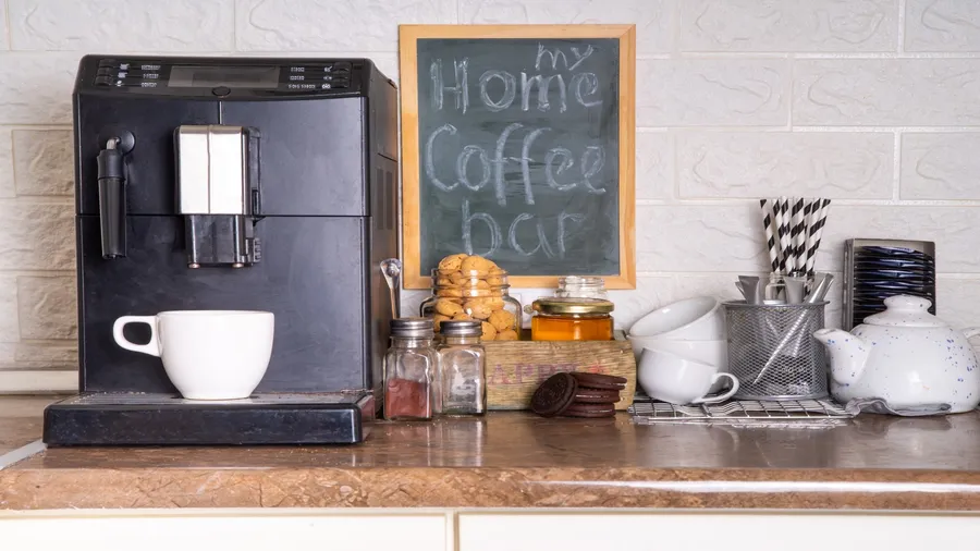 A coffee station with a coffee maker, cups, biscuits, and a chalkboard on a kitchen countertop.