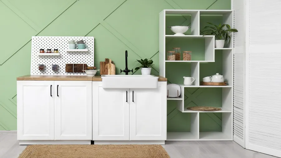 A kitchen interior with white cabinets, wooden countertop, white shelf with plants and jars, rug, and a pegboard in front of a mint green wall.