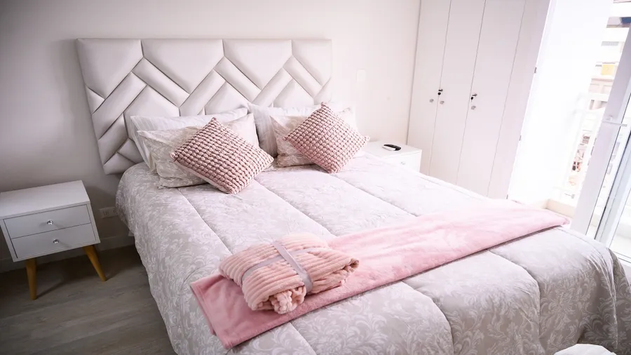 A bedroom interior with a white nightstand, cupboard, and a bed with pillows and upholstered geometric patterned headboard.