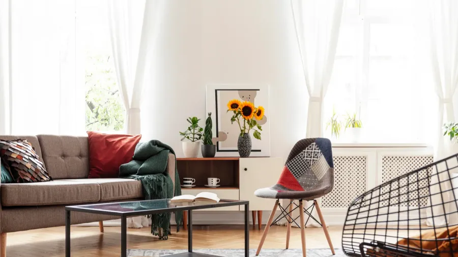 A white living room interior with brown sofa with colorful cushions and throw, coffee table, accent chair, and white sheer curtains on the windows.
