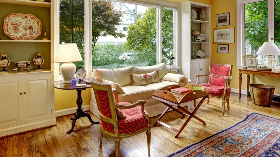 Sitting area with antique furniture. A cozy sitting area with a cream-colored sofa, two patterned red chairs, and a small side table, complemented by large windows letting in natural light and greenery outside.