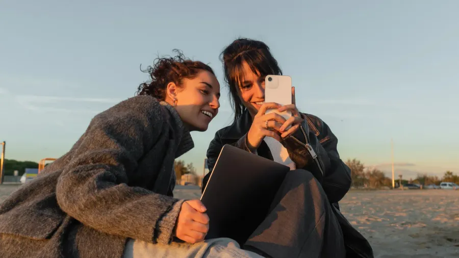 smiling friends using smartphone and laptop on beach in spain