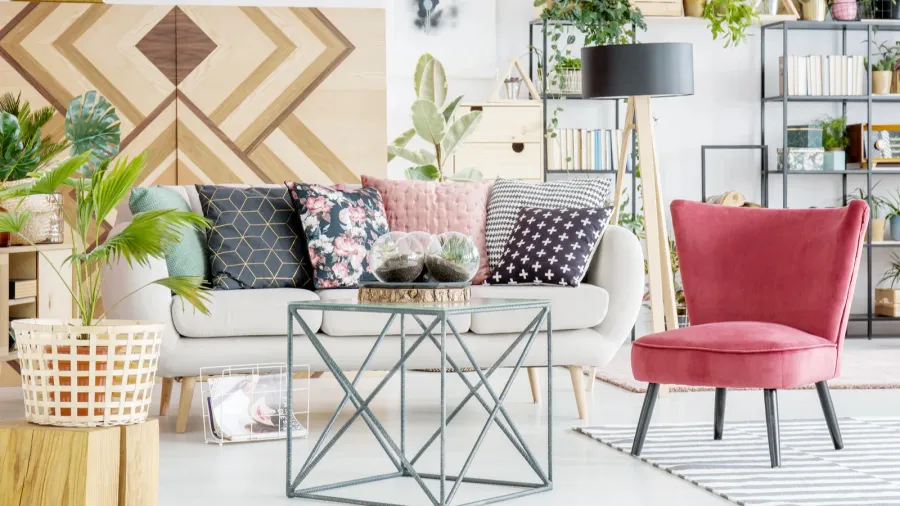 A cozy living room with a white sofa, colorful pillows, plants, and a geometric wall design.