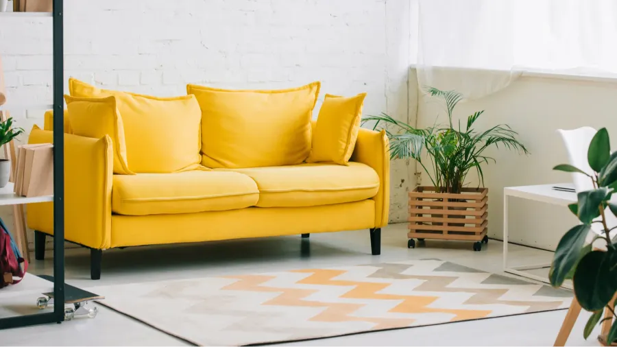 A bright yellow sofa with pillows in a modern room featuring a patterned rug and plants.