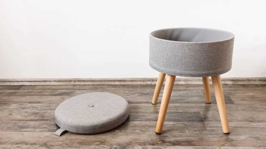A gray ottoman with removable seat for hidden storage on a wooden floor.