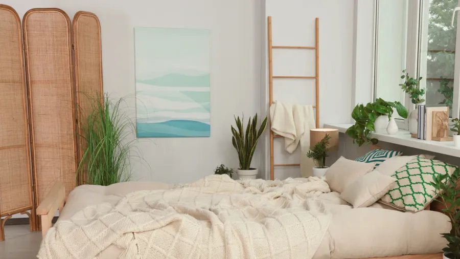 A bright and modern bedroom features a comfortable bed with neutral-toned bedding, a decorative wooden ladder, a woven room divider, and several potted green plants, creating a serene and natural aesthetic.