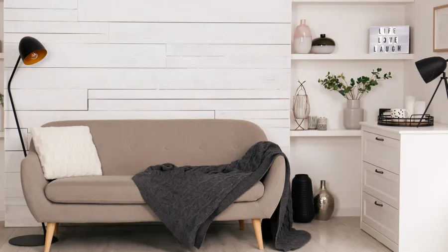 Beige sofa with a white pillow, dark gray throw, black floor lamps, white cabinet with decor, shelves with vases, and a rustic panel wall.