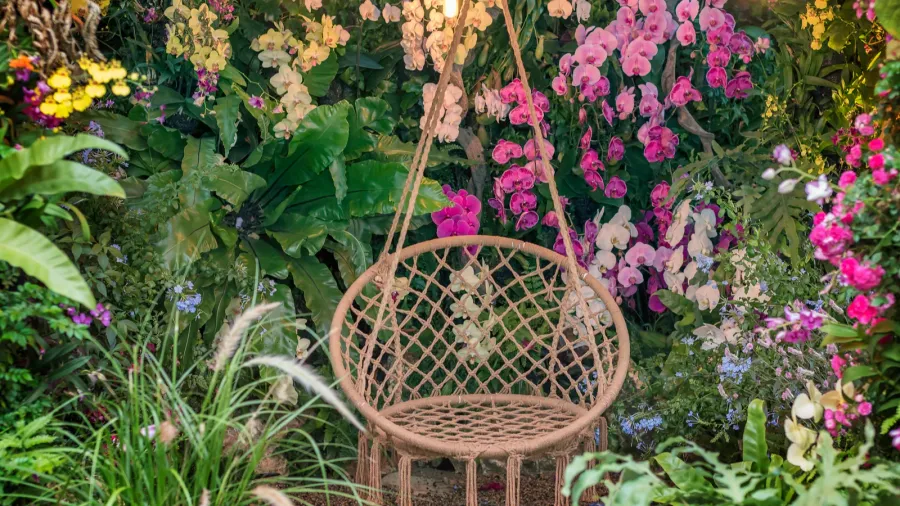 A hanging wicker chair surrounded by vibrant pink, purple, and white flowers in a lush garden.