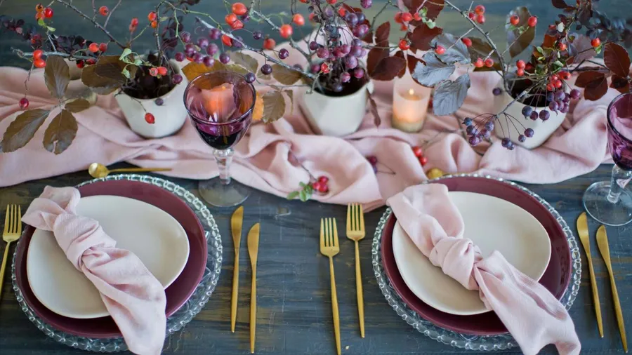 A stylish dining setup with a soft table runner, dinnerware, napkins, small potted plants, and candles.
