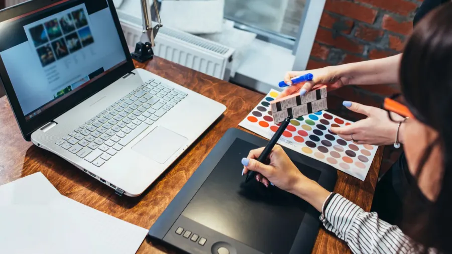 A female interior designer works at a desk with a laptop displaying digital room layouts. She holds a color swatch palette and stylus pen, actively selecting shades while using a graphic tablet. The workspace features a modern setup with a brick wall, desk lamp, and design sketches.