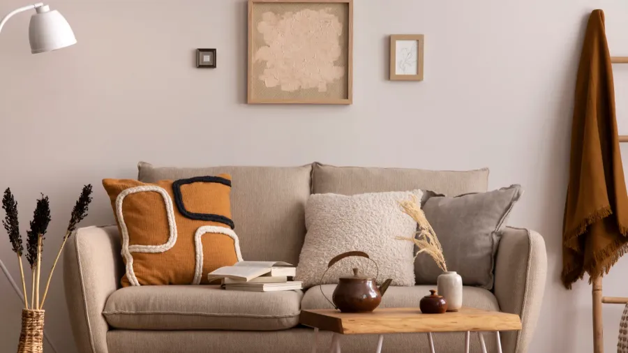 A beige living room interior with sofa with throw pillows and books, wooden coffee table, and framed art on wall.
