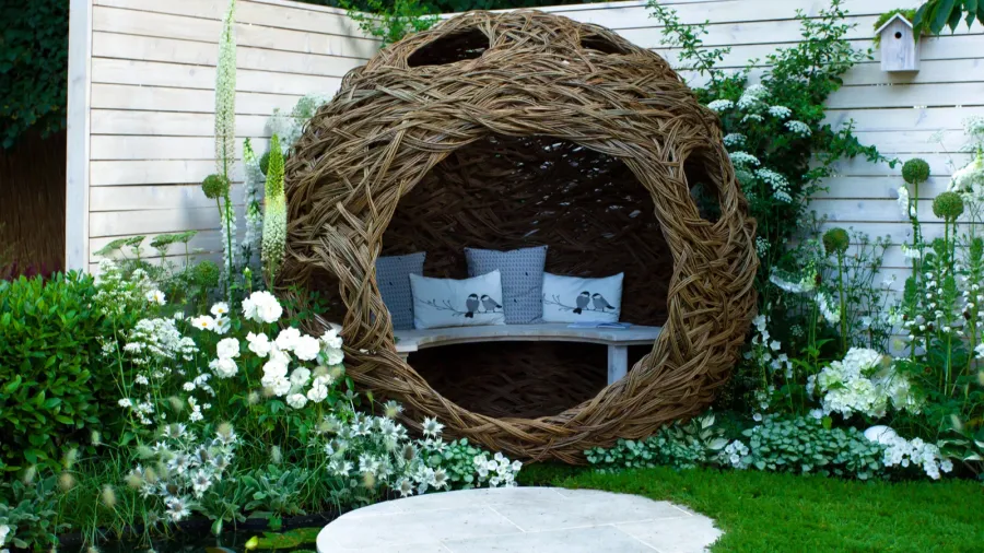 A cozy garden nook with a woven nest-like seat, surrounded by lush greenery and blooming flowers.