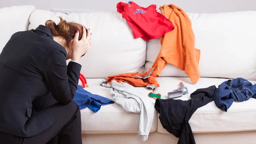 A messy living room with an unhappy woman sitting with hands on her head.