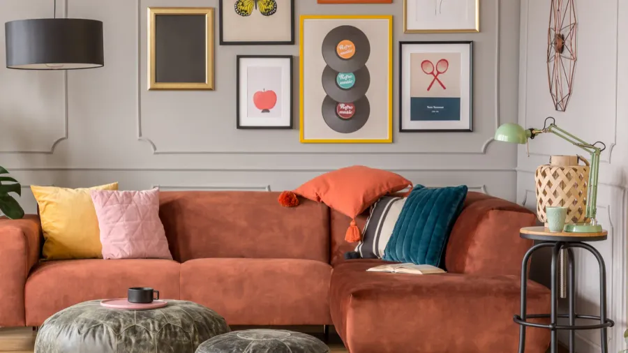 An eclectic living room interior with velvet sectional sofa with colorful throw pillows, poufs, framed artwork, and side table with lamp and woven lantern.