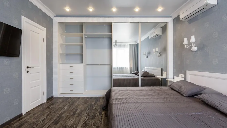 A bedroom interior with a screen on the wall, closet with mirrored doors, and a a cozy bed on a wooden floor.
