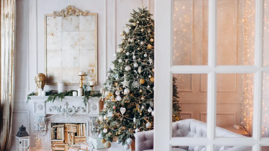 A warm, cozy, and beautiful modern Christmas design living room. White furniture, and decor including console table, sofa, candles, and lanterns. Christmas tree adorned with silver and golden baubles and stars.