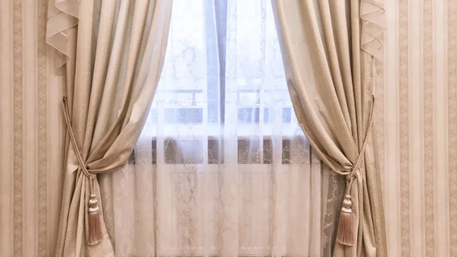 A close-up shot of a window adorned with elegant, light-colored curtains and sheer white inner curtains, tied back with decorative tassels.