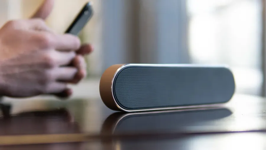 A sleek wireless speaker on a table with a man holding smartphone in his hands in the background.