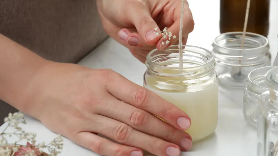 A woman making candle in a glass jar kept on a white table.